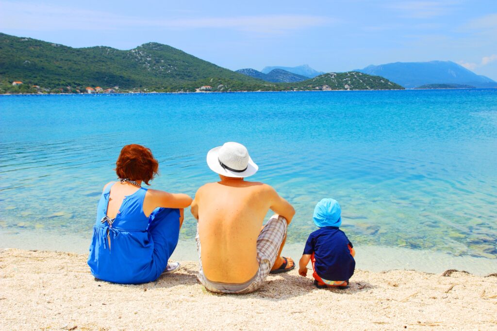 husband and wide sitting on a beach in croatia with their child playing in the sand next to them